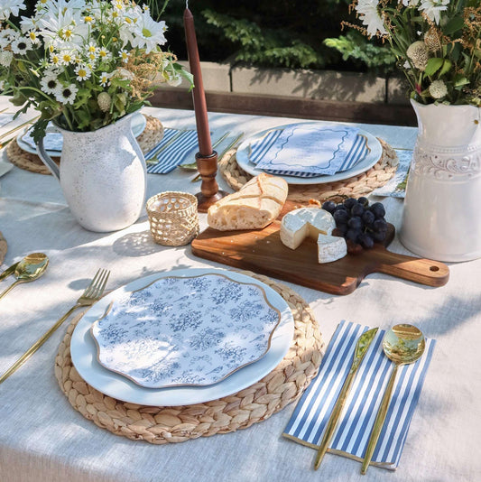 Blue floral toile and blue striped paper napkins on a table set with white plates, gold cutlery, and white flower vases.