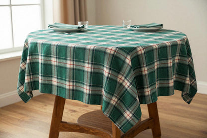 A green plaid tablecloth draped over a round wooden table, styled with folded napkins and glassware in a bright room with a window and wood floor.