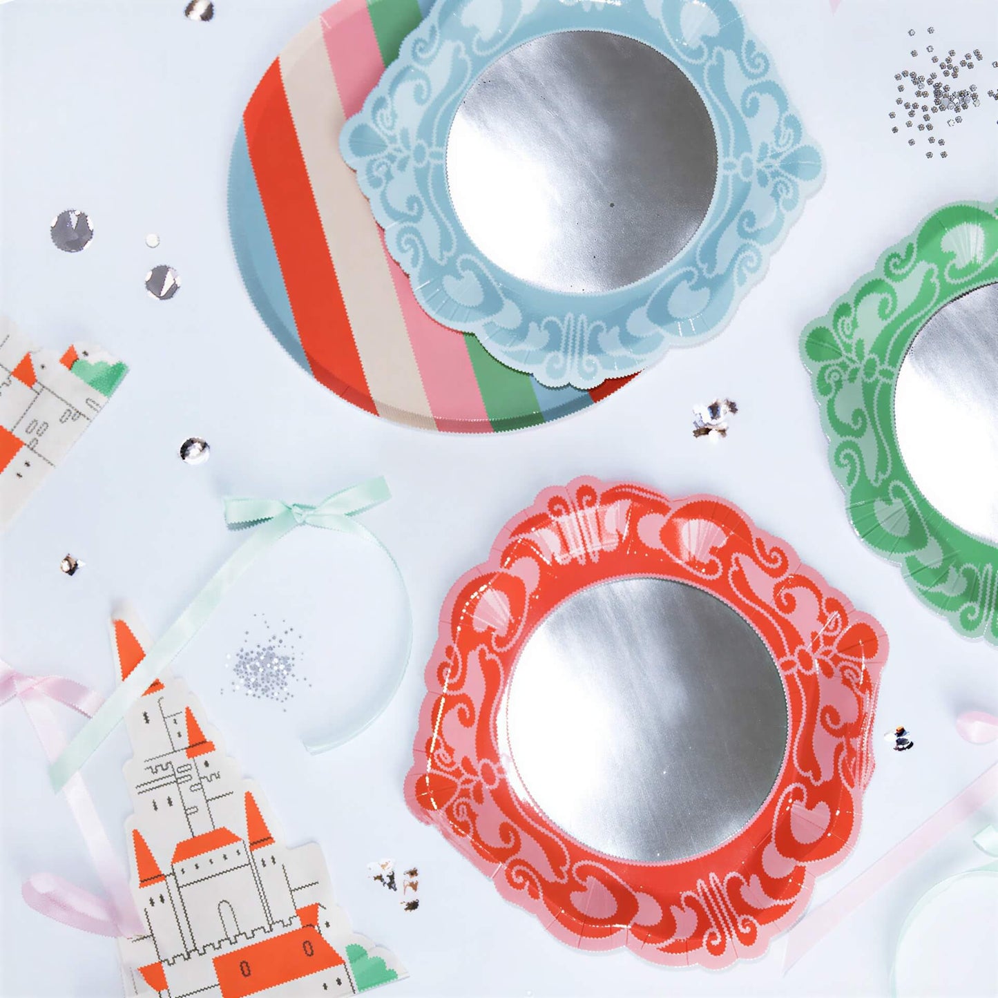 Three round paper plates with silver centers and ornate blue, green, and pink borders near a castle cutout and confetti.