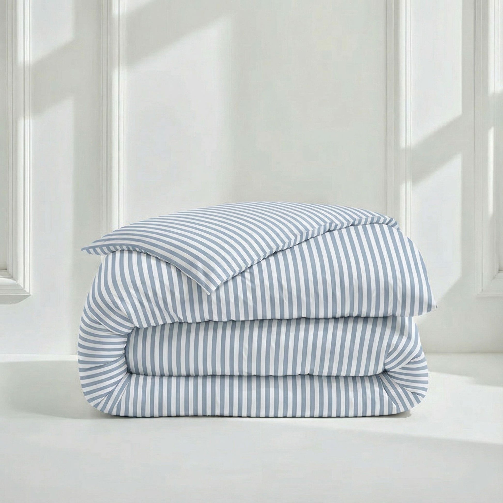 A neatly folded bedding set with a matching pillowcase, featuring light blue and white vertical stripes, displayed on a white surface against a white paneled wall with soft window shadows.
