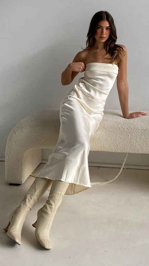 Satin strapless backless midi dress in cream, featuring a side slit, worn by a model sitting on a textured white bench.