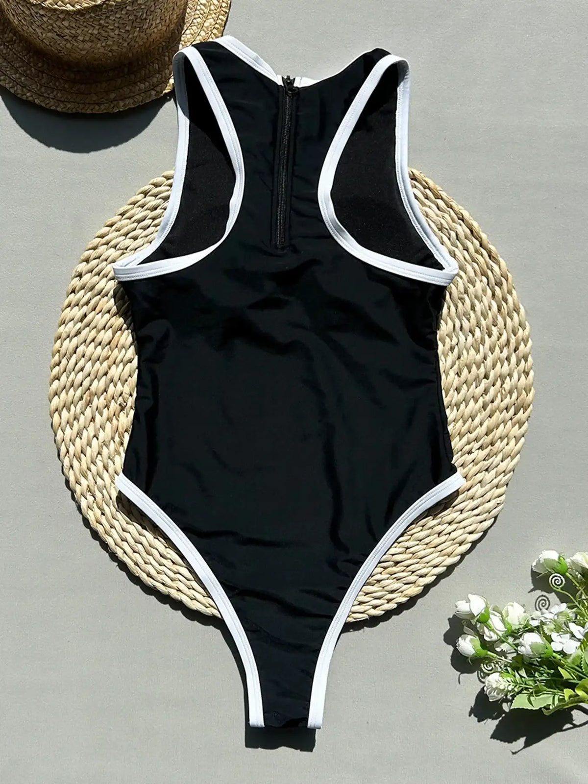 Black swimsuit with white trim on a gray surface with a straw hat and flowers.