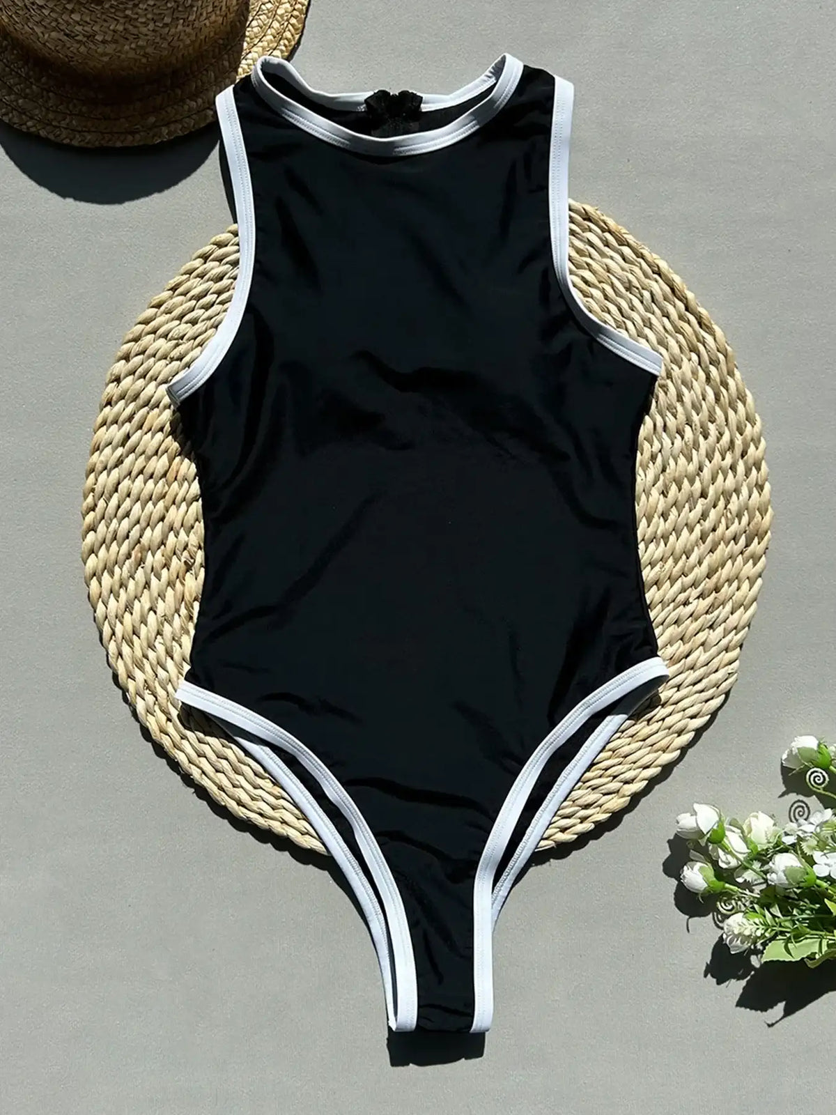 Black swimsuit with white trim on a woven mat with a hat and flowers.