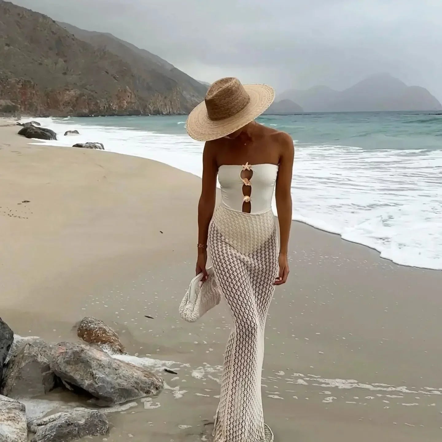 Woman in a strapless white top and fishnet stockings walking on a beach with mountains in the background.