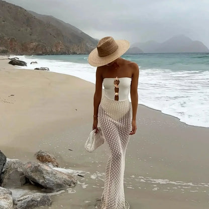 Woman in a strapless white top and fishnet stockings walking on a beach with mountains in the background.
