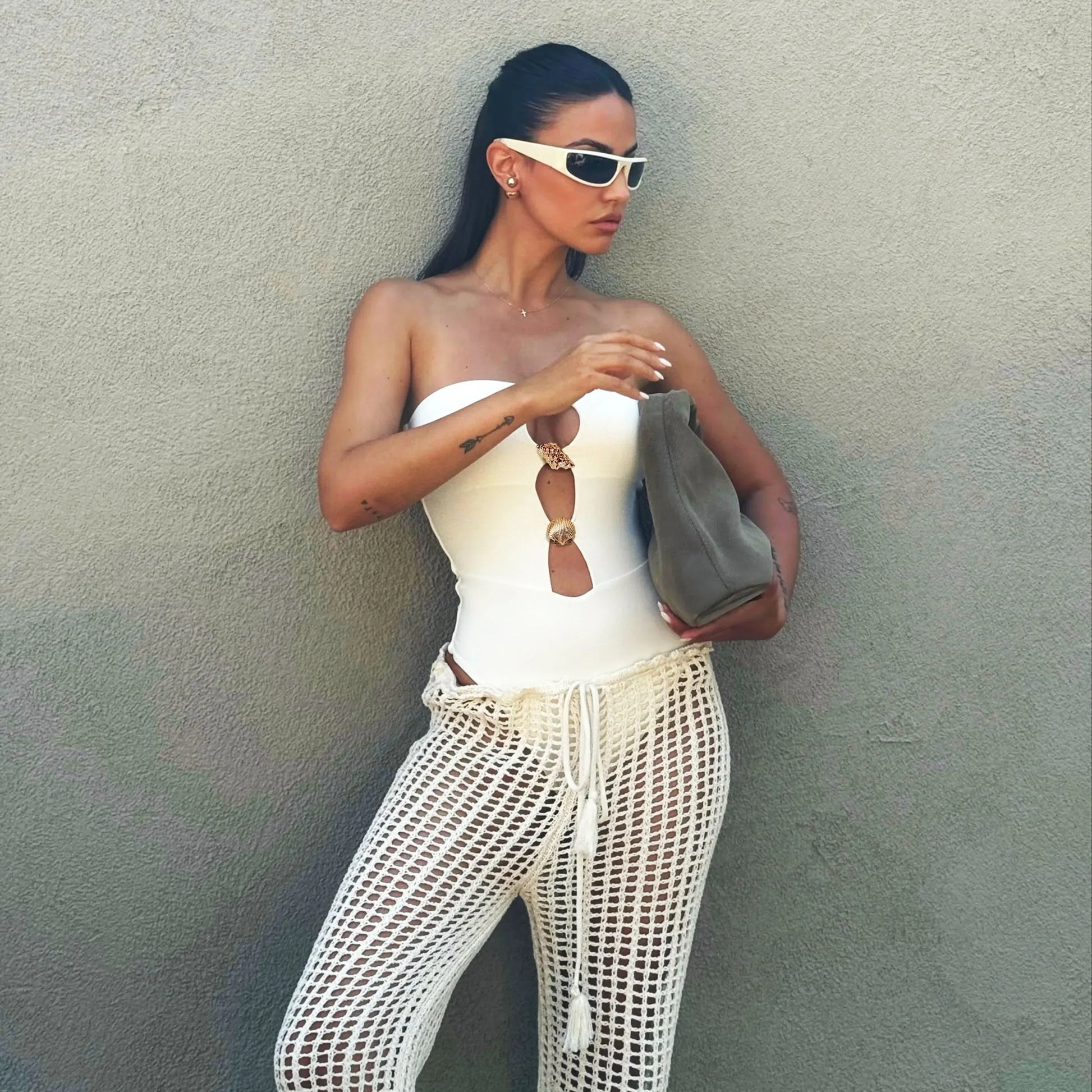 Woman in white strapless top and mesh pants holding a gray clutch against a plain wall.