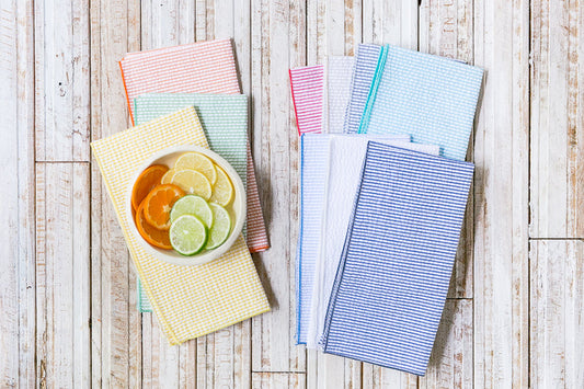 Eight colorful seersucker fabric napkins in various pastel shades arranged on a white wood surface with a bowl of citrus.