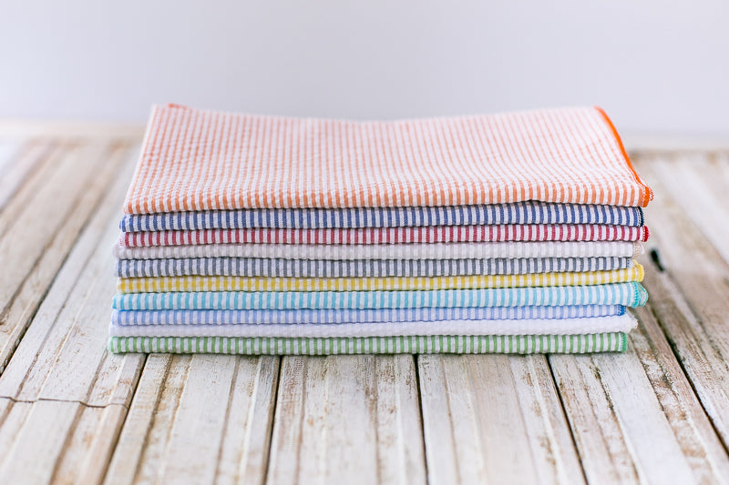 Stack of eight rectangular seersucker fabric napkins in various striped colors on a light wood surface.