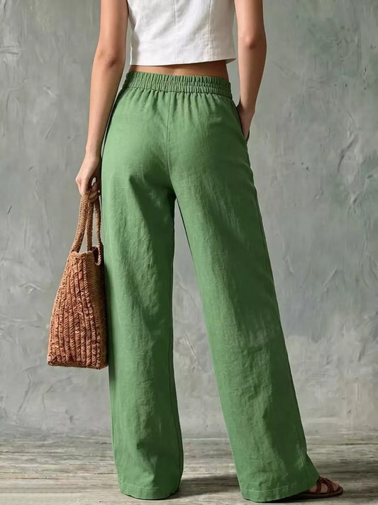 A person wearing loose, wide-leg sage green pants with a white cropped top, holding a woven brown tote bag against a textured gray wall.