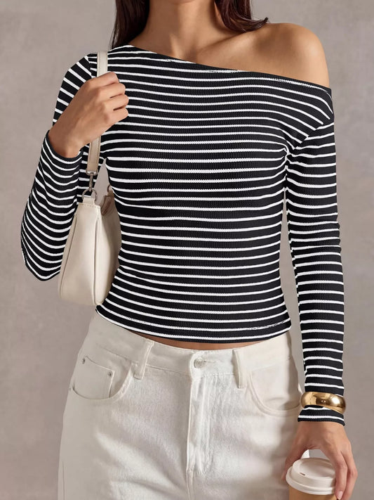 A woman wearing a black and white horizontally striped off-the-shoulder long-sleeve crop top, paired with white jeans, holding a white shoulder bag and a coffee cup against a neutral gray background.