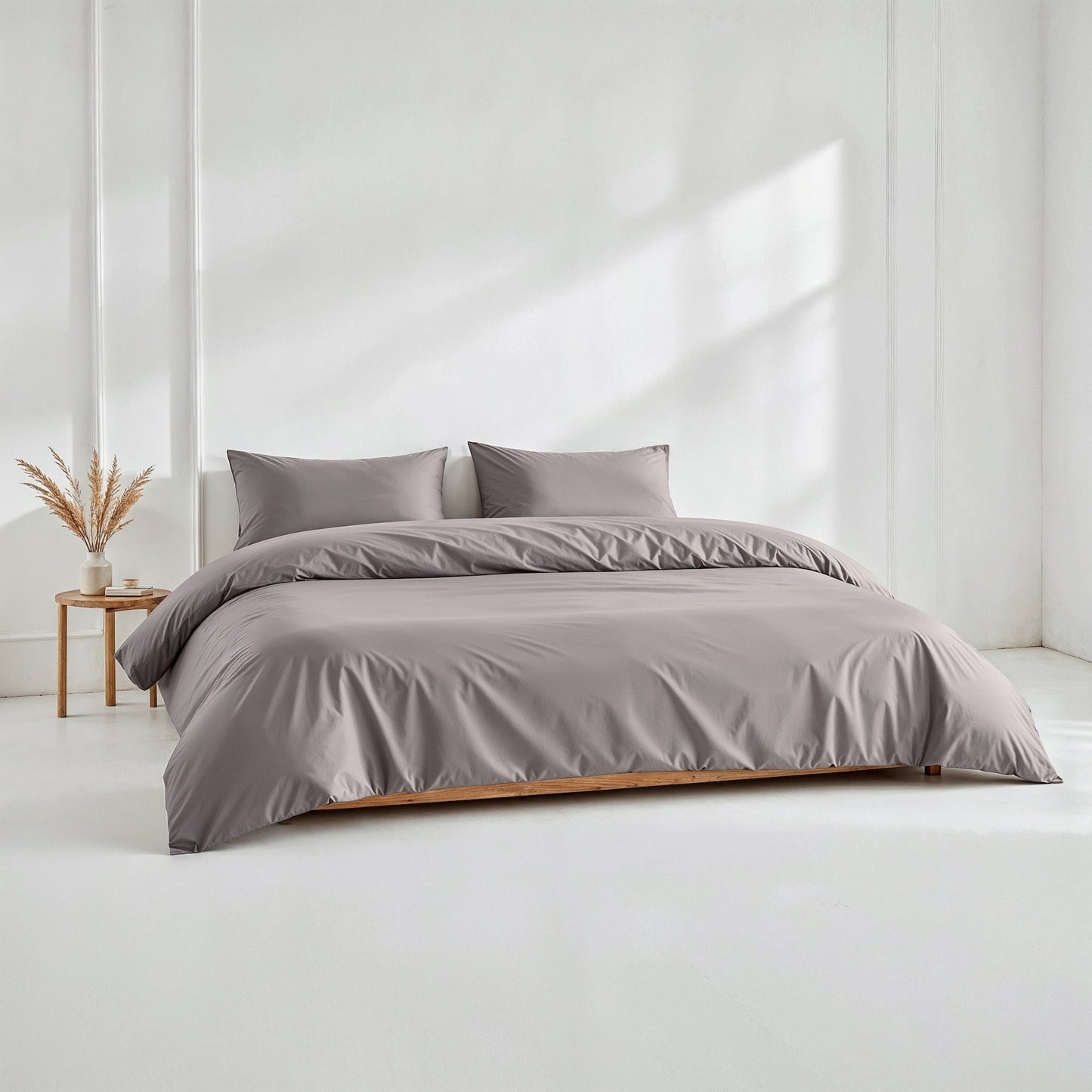 A neatly made bed with a smooth, solid taupe-gray duvet cover and two matching pillowcases, set in a bright, minimalist bedroom with a small wooden side table and dried grass decor.