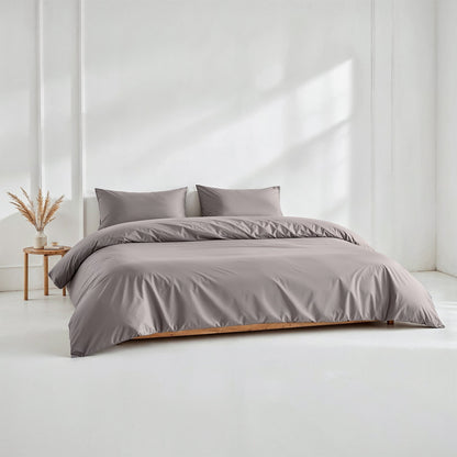 A neatly made bed with a smooth, solid taupe-gray duvet cover and two matching pillowcases, set in a bright, minimalist bedroom with a small wooden side table and dried grass decor.