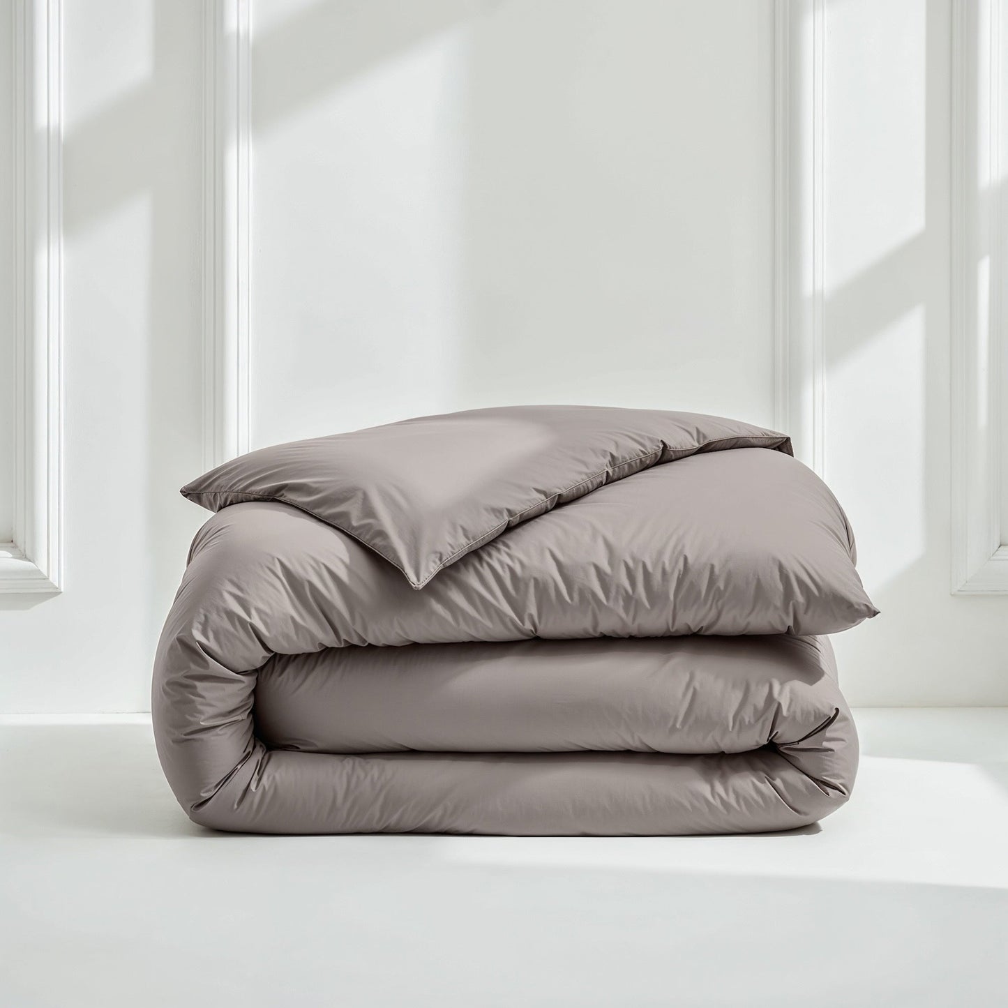 A neatly folded bedding set consisting of a duvet cover and a pillowcase in a solid taupe-gray color, displayed on a white floor against a white paneled wall with soft window light.