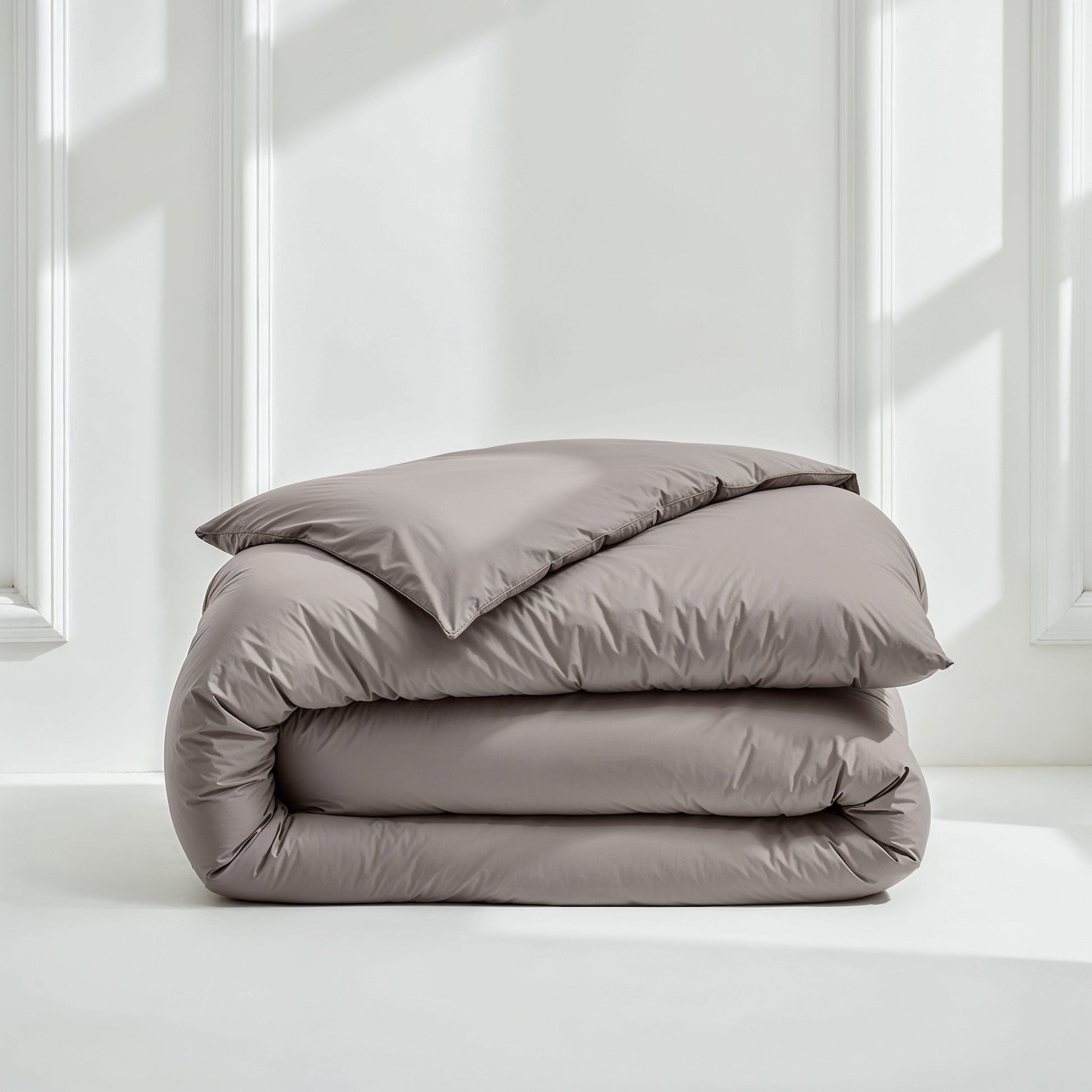 A neatly folded bedding set consisting of a duvet cover and a pillowcase in a solid taupe-gray color, displayed on a white floor against a white paneled wall with soft window light.