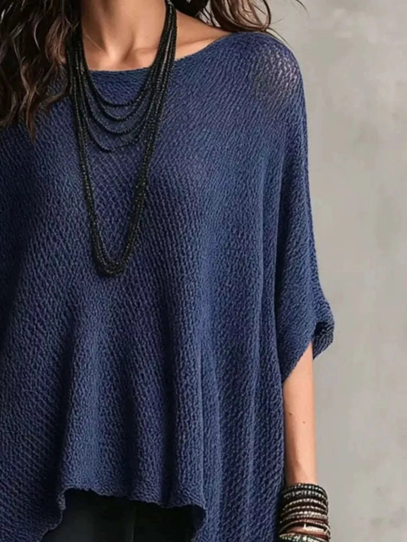 A close-up of a person wearing a loose, textured navy blue knit top with wide short sleeves, layered with multiple long black beaded necklaces and stacked bracelets against a neutral gray background.