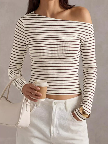 A woman wearing a ribbed off-the-shoulder long-sleeve crop top with thin brown horizontal stripes on an off-white base, paired with white pants and holding a coffee cup.