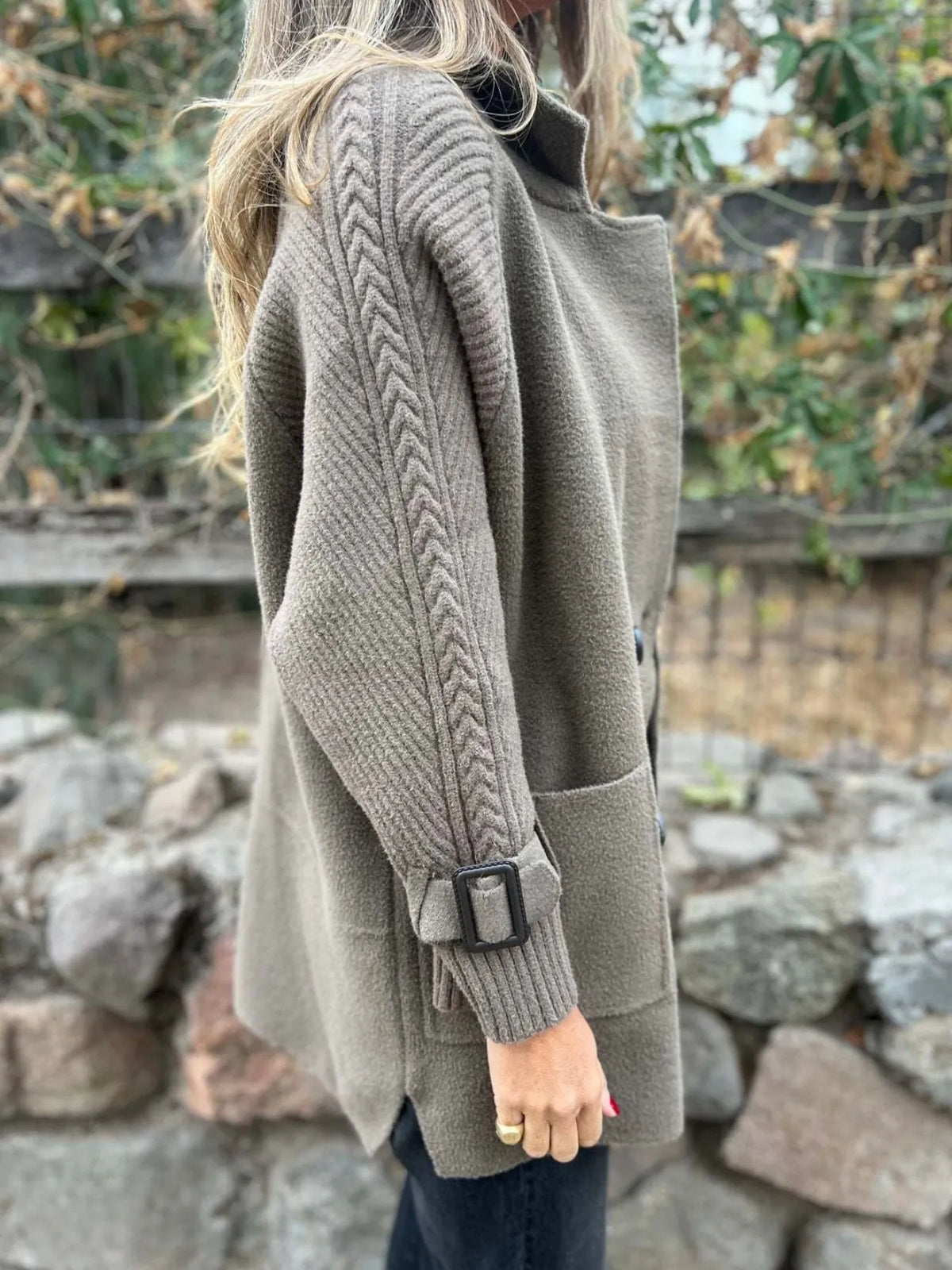 Model wearing a mocha double-breasted coat with cable-knit sleeves, buckled cuffs, wide lapels and front pockets.