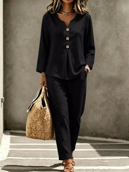 A woman wearing a black two-piece linen set consisting of a long-sleeve button-up top and matching wide-leg trousers, holding a woven straw tote bag against a textured gray concrete wall.