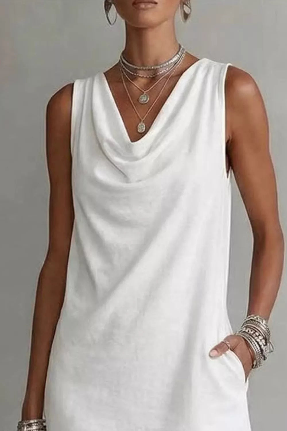 Woman wearing a white sleeveless dress with layered necklaces and bracelets on a gray background