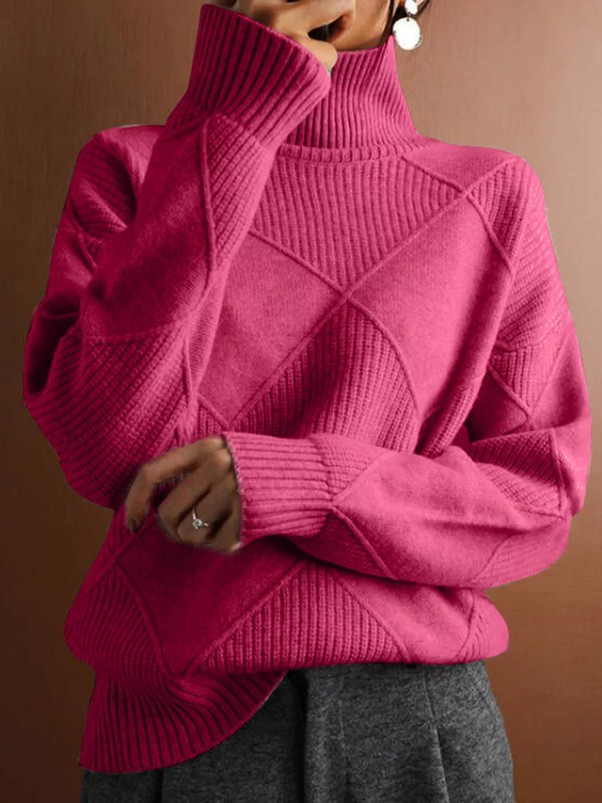 A close-up of a person wearing a bright pink ribbed turtleneck sweater with diamond-shaped knit patterns on the front, paired with gray pants against a warm brown background.