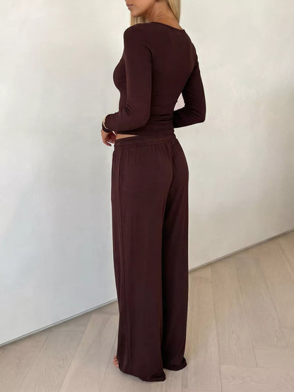 A woman wearing a matching dark brown long-sleeve top and wide-leg pants set, shown from a side-back angle in a minimalist room with a white wall and light wood floor.