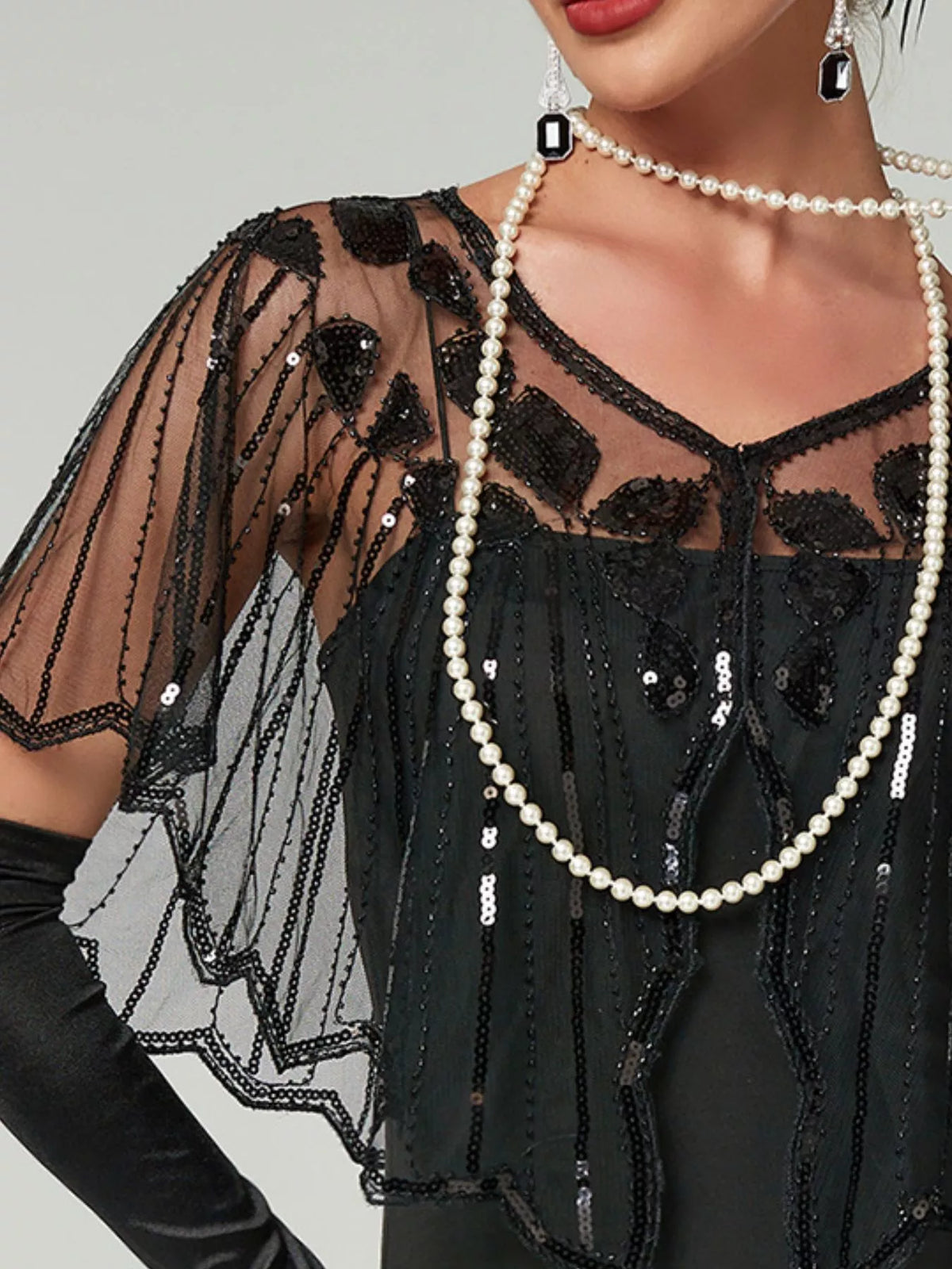 Model wearing a black sheer sequin capelet with a shallow V-neck, floral embellishments and scalloped edges over a black dress