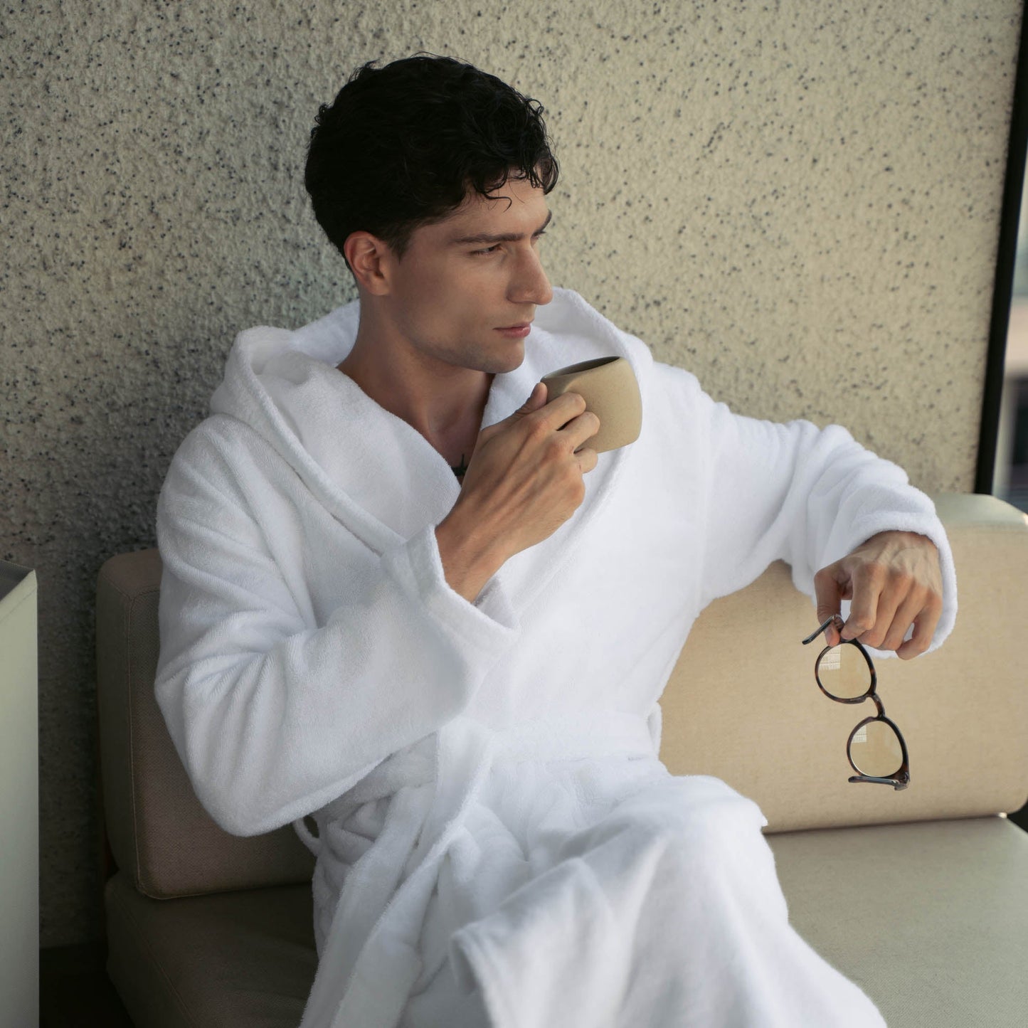 Man in a white robe holding a cup and glasses, sitting on a couch.