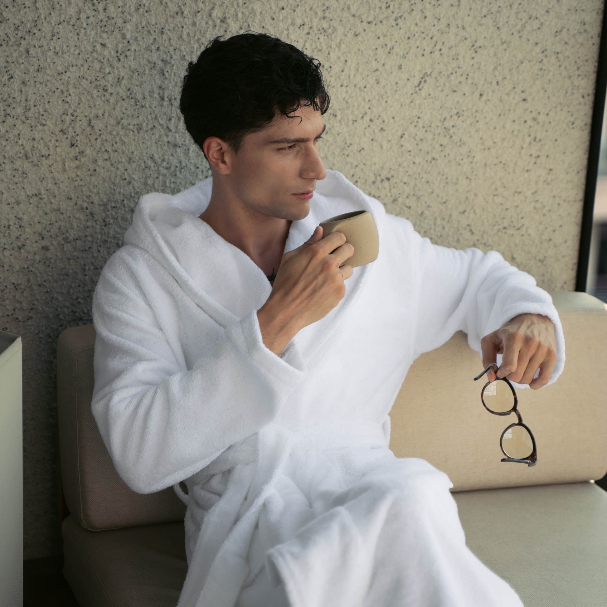 Man in a white robe holding a cup and glasses, sitting on a couch.