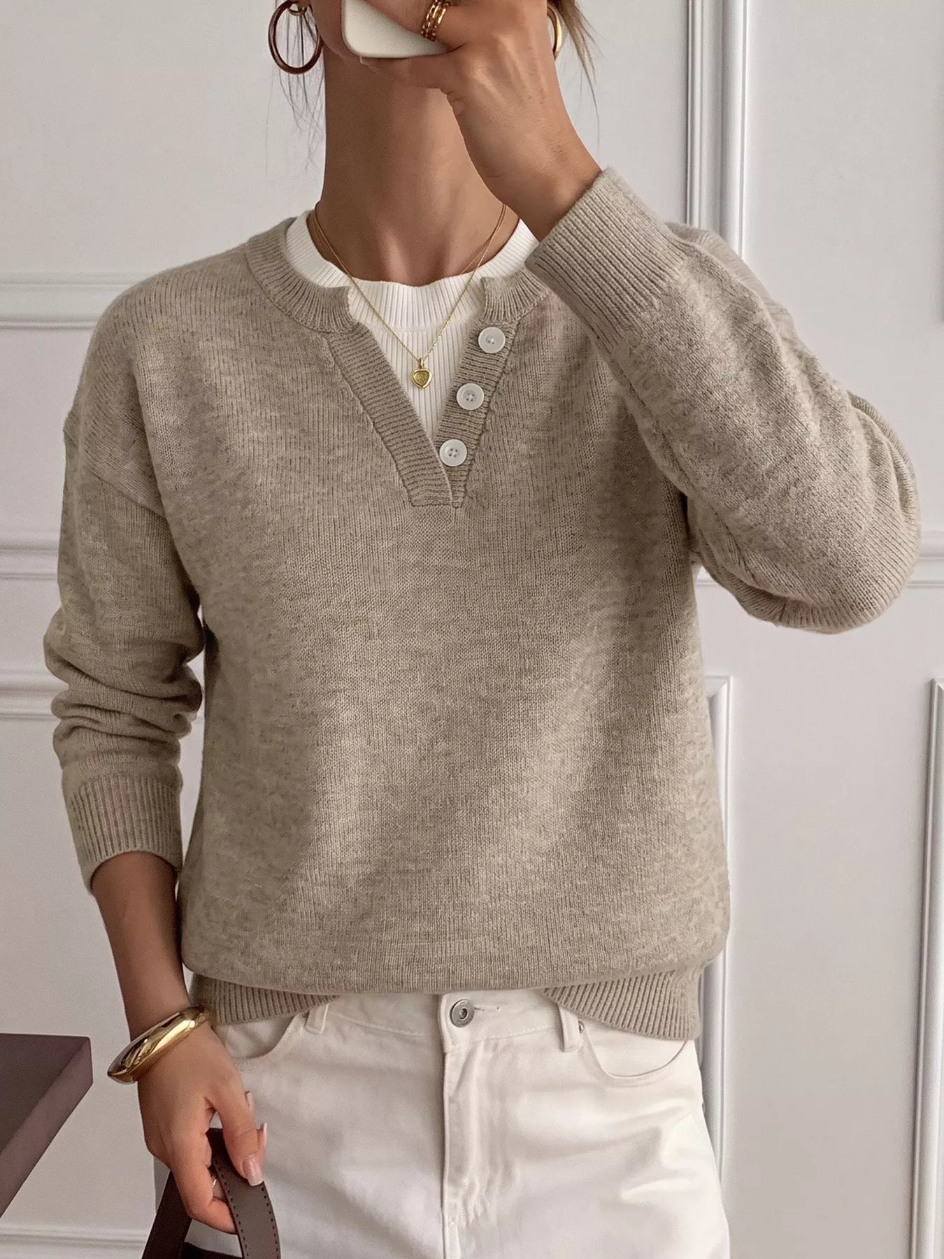 A woman wearing a light beige knit sweater with a split V-neck and three white buttons, layered over a white ribbed top and paired with white pants.