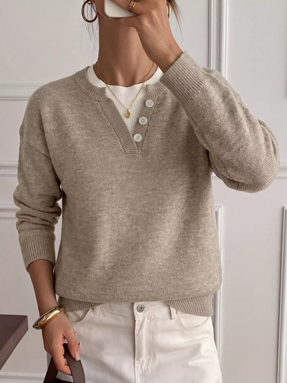 A woman wearing a light beige knit sweater with a split V-neck and three white buttons, layered over a white ribbed top and paired with white pants.