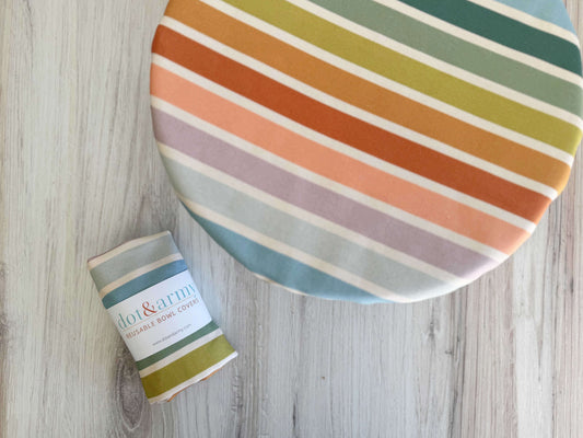 Round fabric bowl cover with multicolor stripes and a rolled cover with a dot & army label on a light wood surface.
