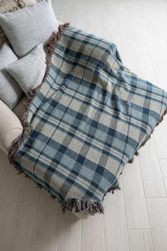 A plaid throw blanket draped over a white chair, featuring a blue, gray, and white tartan pattern with fringed edges, set against a light gray brick wall background.