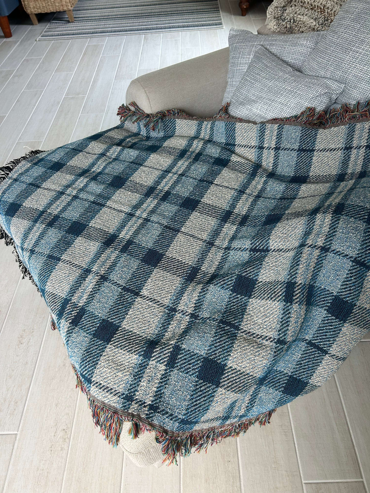 A plaid throw blanket draped over a white chair, featuring a blue, gray, and white tartan pattern with fringed edges, set against a light gray brick wall background.