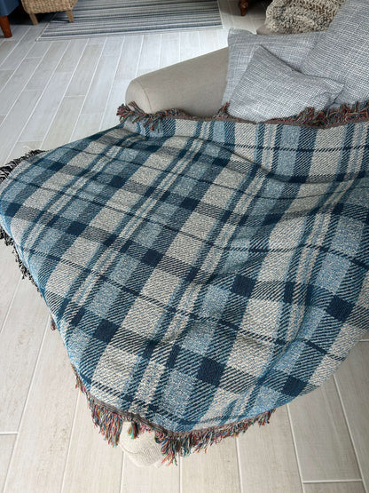 A plaid throw blanket draped over a white chair, featuring a blue, gray, and white tartan pattern with fringed edges, set against a light gray brick wall background.