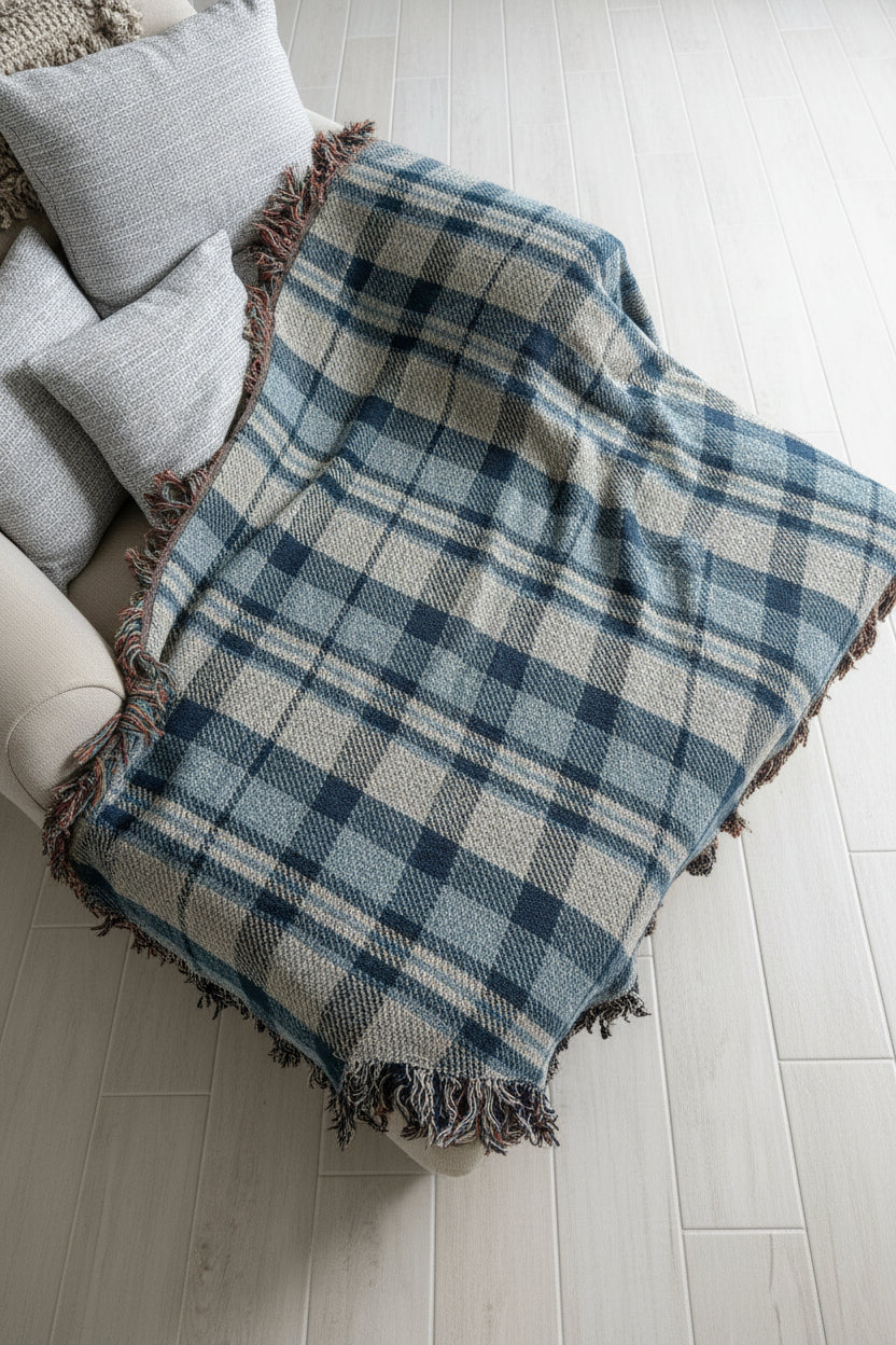 A plaid throw blanket draped over a white chair, featuring a blue, gray, and white tartan pattern with fringed edges, set against a light gray brick wall background.