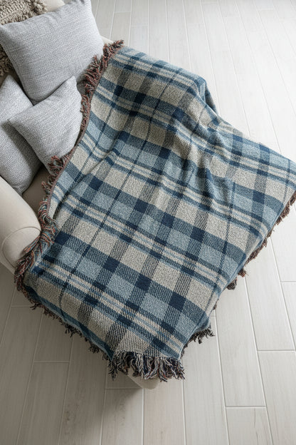 A plaid throw blanket draped over a white chair, featuring a blue, gray, and white tartan pattern with fringed edges, set against a light gray brick wall background.