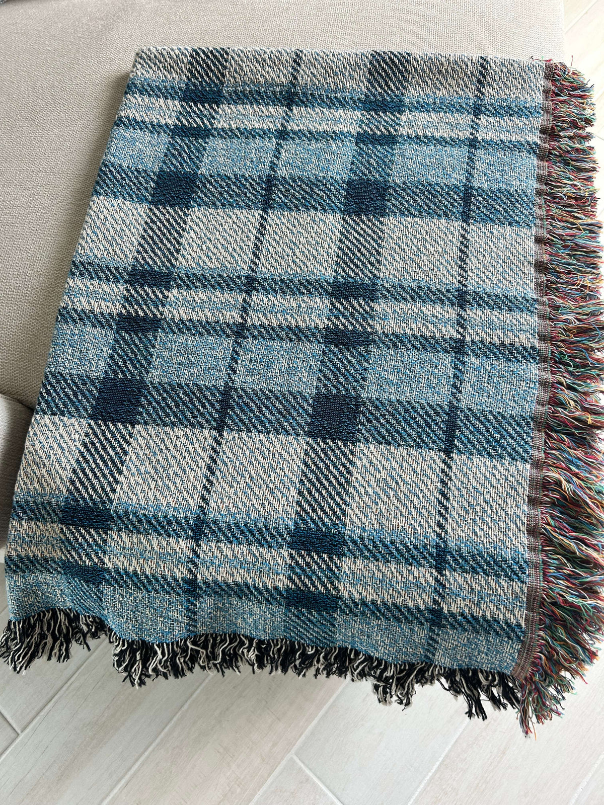 A woven plaid throw blanket in shades of blue, gray, and white, featuring multicolored fringe on one side, laid out on a light-colored sofa over a light wood floor.
