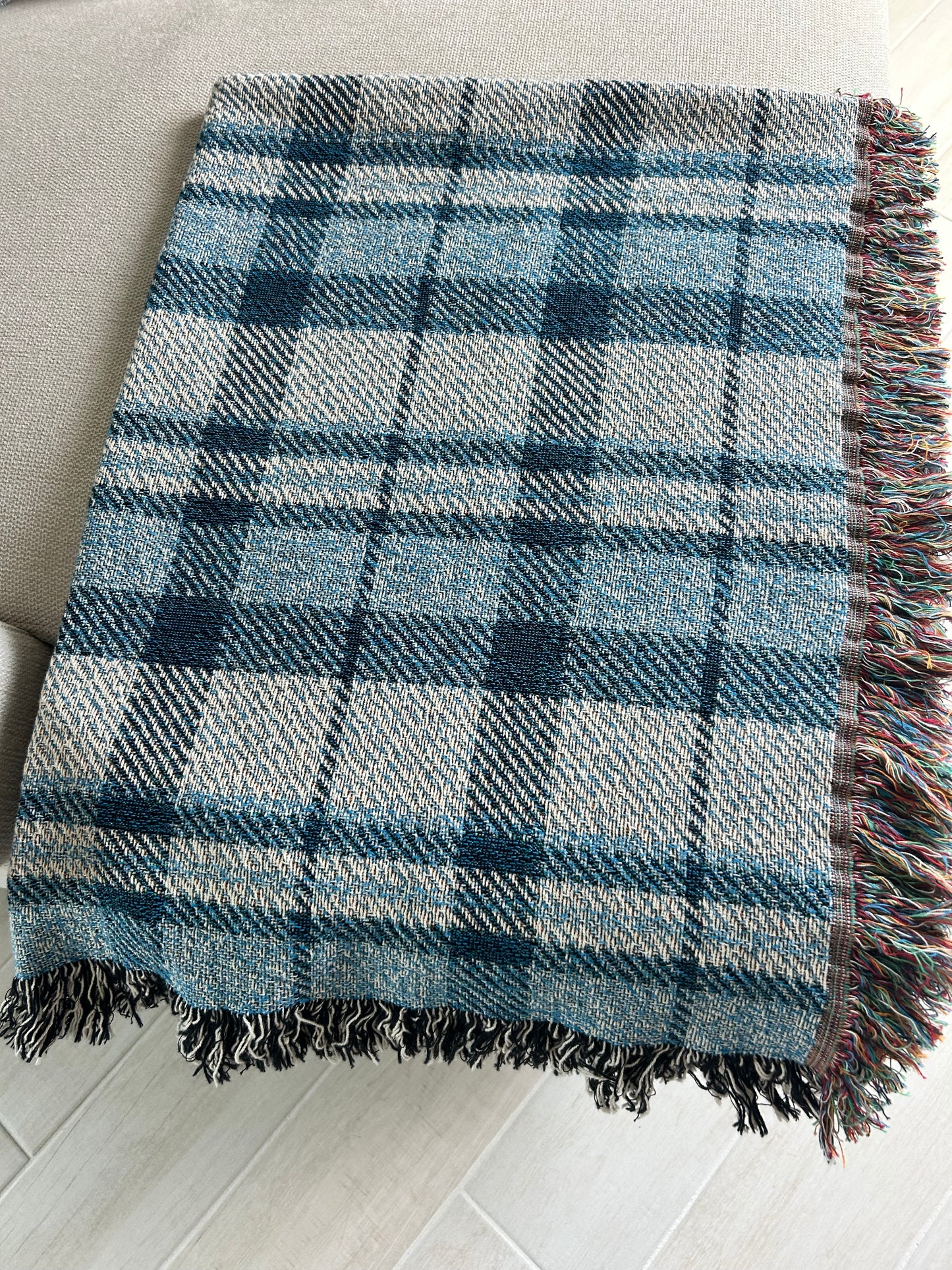 A woven plaid throw blanket in shades of blue, gray, and white, featuring multicolored fringe on one side, laid out on a light-colored sofa over a light wood floor.