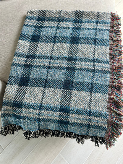 A woven plaid throw blanket in shades of blue, gray, and white, featuring multicolored fringe on one side, laid out on a light-colored sofa over a light wood floor.