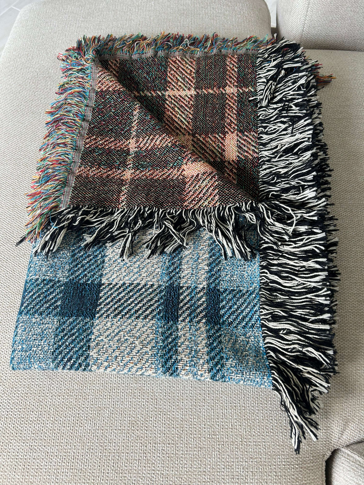 A folded plaid throw blanket with fringed edges, laid on a light beige upholstered chair. One half shows a dark brown, black, and dusty pink plaid pattern, while the other half shows a blue, gray, and white plaid pattern.