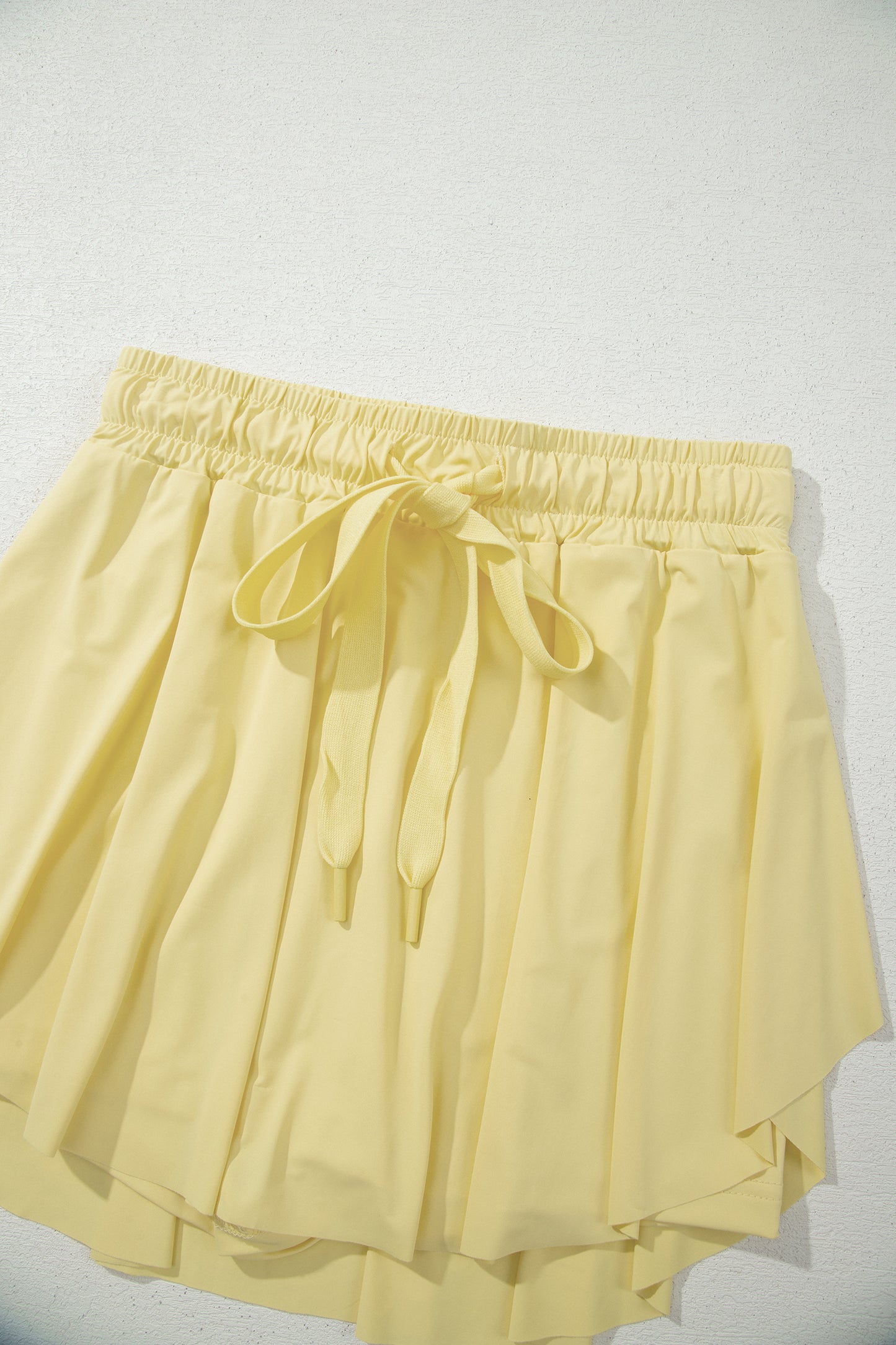 A pair of light yellow high-low pleated shorts with an elastic waistband and a matching drawstring bow, shown against a plain light background.