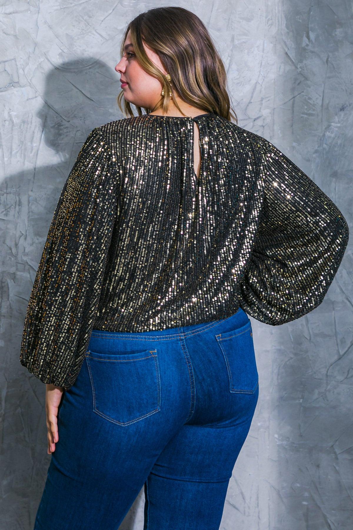 Woman wearing a sparkly black top and blue jeans against a gray textured wall.