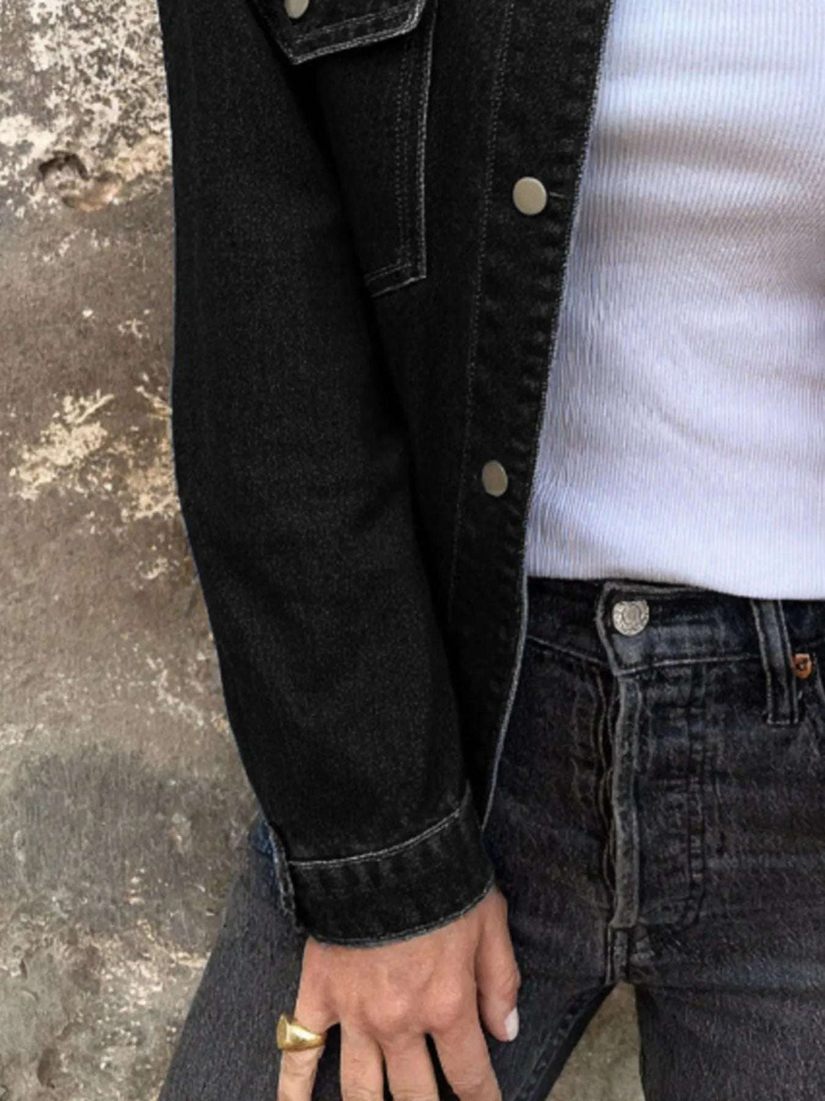 Person wearing a black denim jacket and jeans against a textured wall.