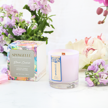 Black Orchid scented candle in a clear glass holder, with a decorative box and surrounding flowers.