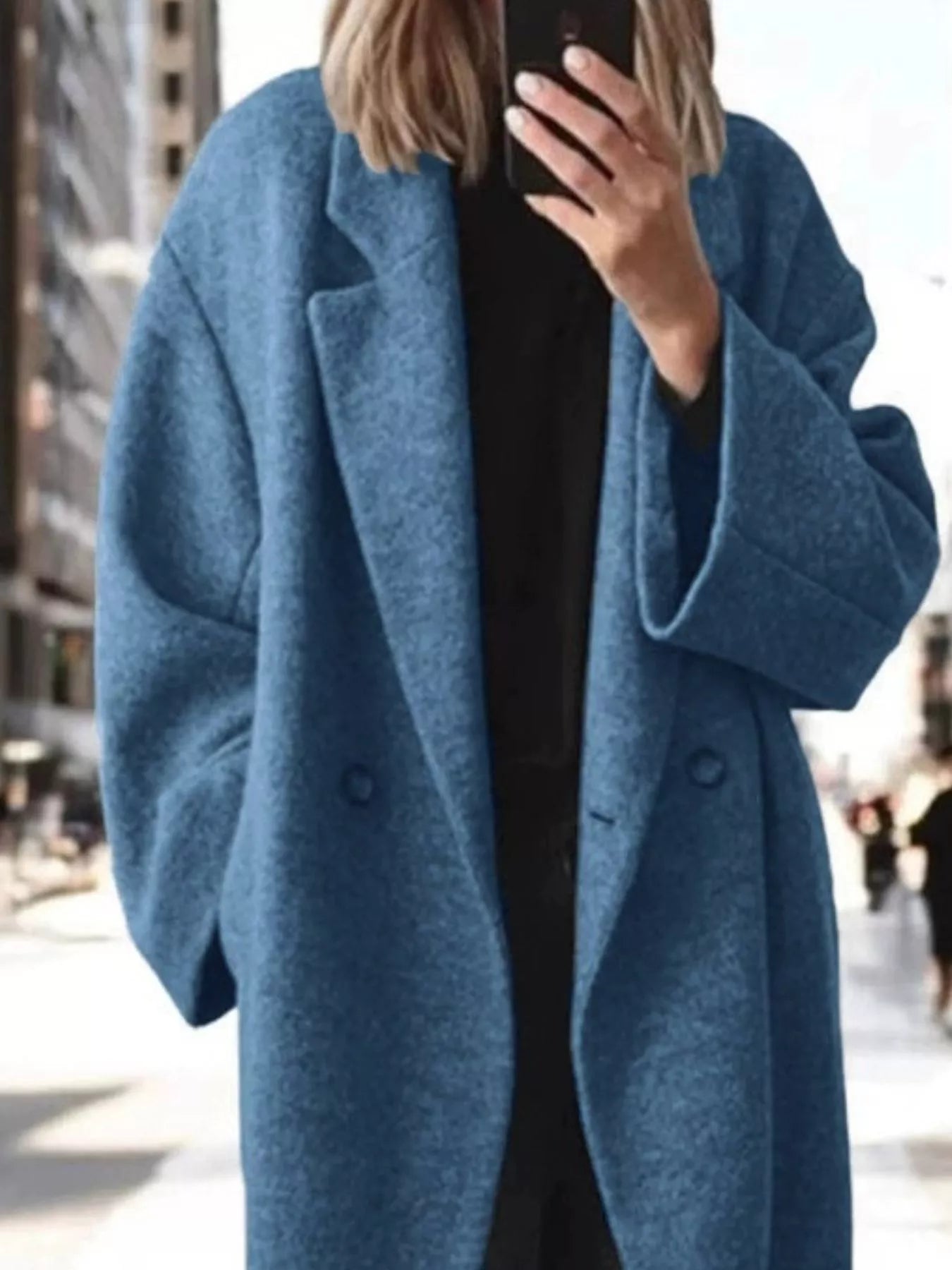 A person taking a mirror selfie wearing a textured, heathered blue double-breasted coat over a black top, set against a blurred city street background.