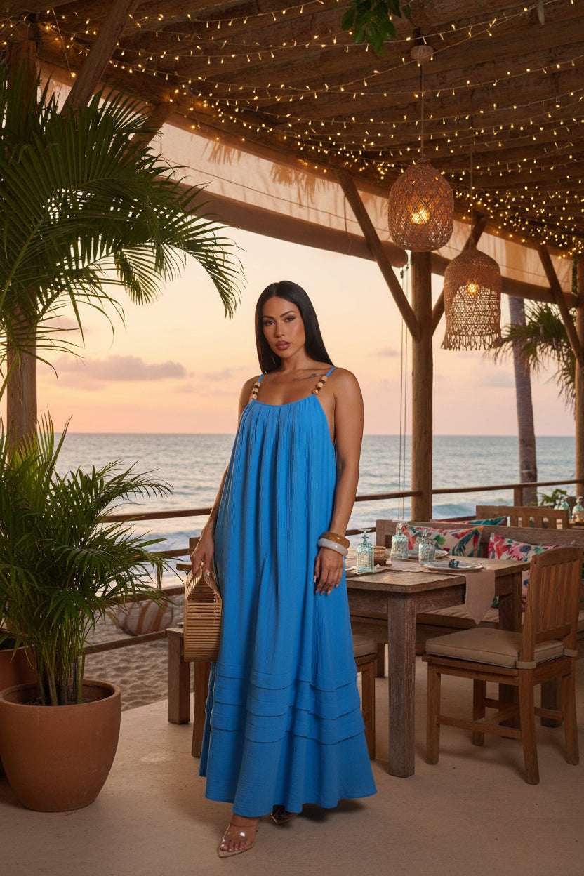 Woman wearing a blue dress holding a woven bag at a beachside restaurant with the ocean in the background.