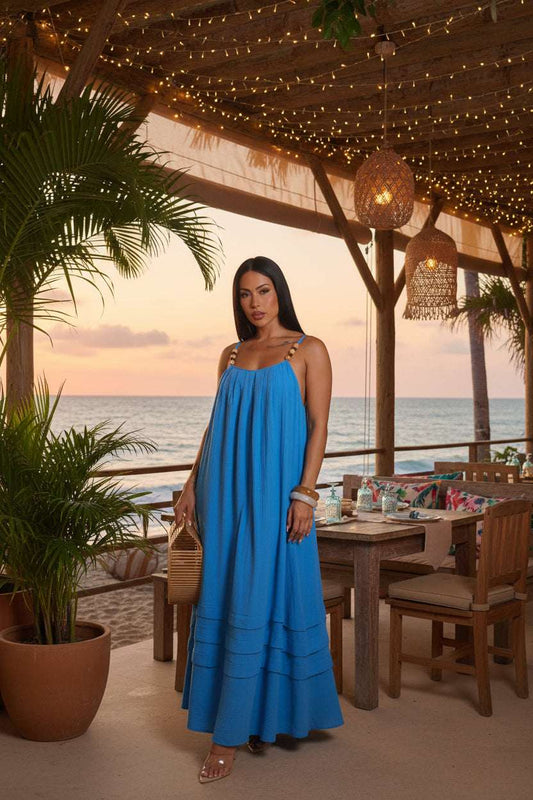 Woman wearing a blue dress holding a woven bag at a beachside restaurant with the ocean in the background.