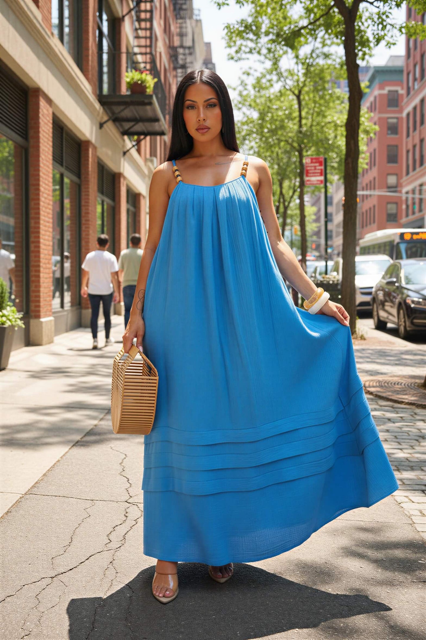 Woman wearing a blue dress holding a woven bag on a city street.