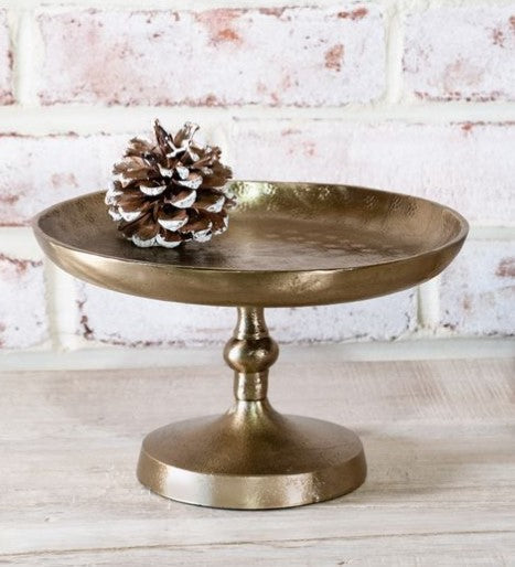 Bronze cake stand with a pine cone on a light wooden surface and white brick wall background