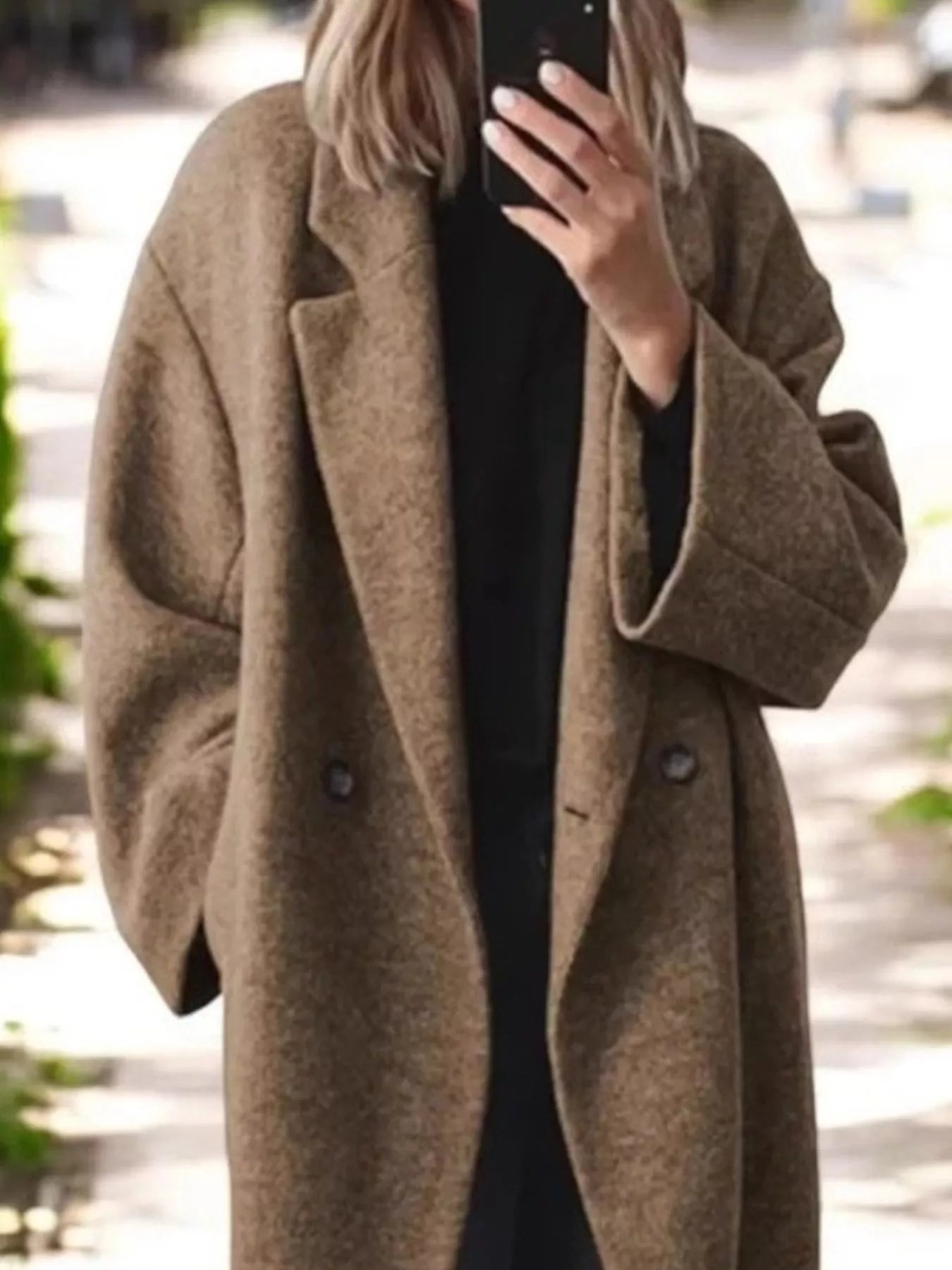 A person outdoors wearing an oversized brown wool-blend coat over a black outfit, holding a phone to take a mirror selfie.